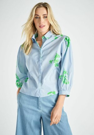Vrouw met lang blond haar draagt een lichtblauw shirt met groene bloempatronen en lichtblauwe broek met wijde pijpen, staand met één hand in de zak.