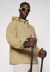 Beige lightweight jacket with a hood, elastic cuffs. Model holds trekking poles. Accessories include light-colored sunglasses. Smooth texture.