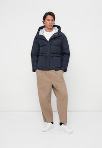 Marineblauwe pufferjack met capuchon, voorzien van een ritssluiting en twee voorzakken, gedragen met beige wijde broek en witte sneakers.
