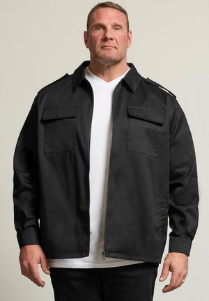 Homme portant une veste noire zippée avec poches à rabat sur la poitrine par-dessus une chemise blanche, debout devant un fond clair uni.