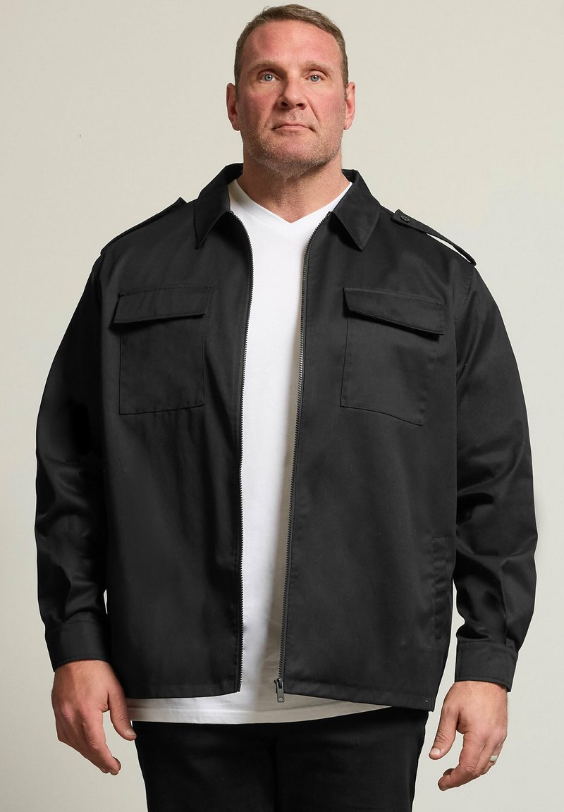 Homme portant une veste noire zippée avec poches à rabat sur la poitrine par-dessus une chemise blanche, debout devant un fond clair uni.