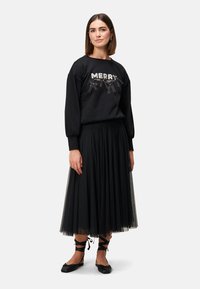 Czarna bluza z napisem "MERRY" wykonanym z cekinów i wstążek, w połączeniu z czarną, plisowaną spódnicą z tiulu oraz czarnymi balerinkami z wiązaniami na kostkach.