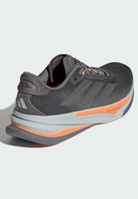 Scarpa sportiva grigia con tomaia texture, tre strisce grigie, dettagli arancioni e suola ammortizzata con scanalature distintive.