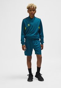 Tealfärgad sportoverall med halvlång dragkedja, svarta axeldetaljer och gul märkning. Kombinerad med tealfärgade shorts och svarta sportskor.