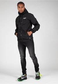 Giacca leggera nera con cappuccio, chiusura a zip e polsini elasticizzati, abbinata a jeans in denim scuri e sneakers nere con accenti verdi.