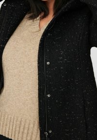 Chaqueta negra texturizada con cierre de cremallera y botones de presión, combinada con un suéter beige que presenta un dobladillo acanalado y un sutil diseño de escote en V.