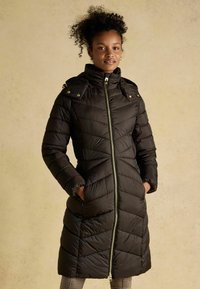 Čierna páperová bunda s vysokým golierom, zipsom a vzorom vo forme šípok. Obsahuje dva bočné vrecká a úzky, po kolená dlhý strih.