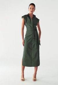 Abito verde oliva con maniche corte, vita svasata, bottoni frontali, cintura abbinata e spacco laterale, indossato con sandali beige con cinturini.