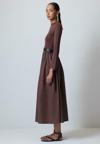 Brun långärmad klänning med en figurnära överdel och en flödande kjol, kombinerad med ett svart bälte. Modellen bär platta svarta sandaler.