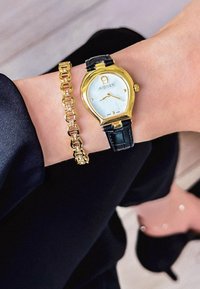 Reloj Aigner dorado con forma de lágrima, esfera blanca y correa de cuero negro, combinado con una pulsera de eslabones dorados en una muñeca.