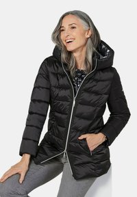 Schwarze Pufferjacke mit Kapuze, mit quiltetem Design, glänzender Oberfläche, silbernen Reißverschlüssen und seitlichen Taschen. Getragen über einem gemusterten Oberteil.