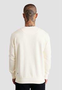 Sudadera de color crema hecha de tela suave, con cuello redondo, puños de canalé y un pequeño acento de logo cerca del dobladillo.