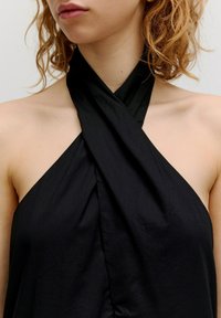 Zwarte haltertop van gladde stof; heeft een gedraaide design bij de nek en een losse silhouet. Blootgestelde schouders en sleutelbeen.