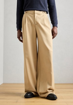 Beige wide-leg trousers with a smooth texture, front pleats, and belt loops, paired with black shoes on a wooden floor.