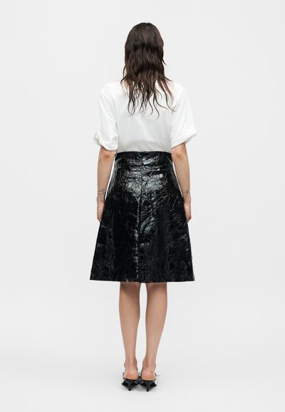 Γυναίκα με κυματιστά καστανά μαλλιά που φοράει λευκό μπλουζάκι με κοντά μανίκια, βγαλμένο μέσα σε μια γυαλιστερή μαύρη φούστα μέχρι το γόνατο και μαύρα παπούτσια με τακούνια, στραμμένη από πίσω.