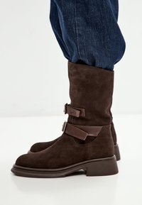 Botines de ante marrón con punta redondeada, que presentan dos tiras de cuero marrón con hebillas de metal y un tacón grueso.