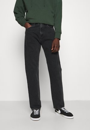 Person iført mørkegrå lige jeans, grøn hættetrøje og sorte sneakers med hvide såler og snørebånd, står med den ene hånd i lommen.