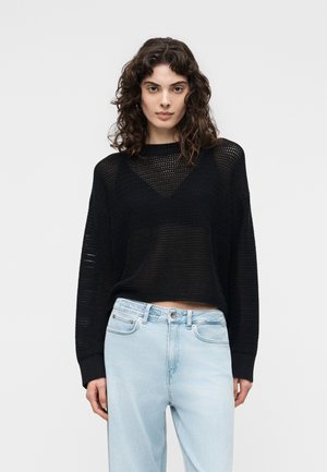 Kvinde med krøllet mørkt hår iført en sort mesh-langærmet trøje over en sort top og lyseblå højtaljede jeans, stående neutralt.