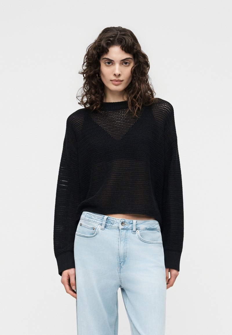 Femme aux cheveux bouclés et foncés, portant un pull noir en maille à manches longues par-dessus un haut noir et un jean taille haute bleu clair, debout en position neutre.