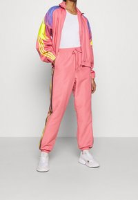 Rosa träningsoverall med en zip-up jacka som har färgglada detaljer. Byxorna har dragsko i midjan och sidostrimmor med gula och svarta detaljer. Vita sneakers.