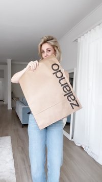 Rjava papirnata nakupovalna vrečka z velikim črnim napisom "zalando", ki jo drži oseba v svetlo modrih kavbojkah, v svetlem notranjem prostoru.