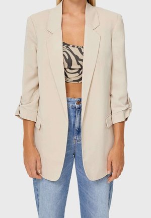 Vrouw draagt een beige blazer over een zwart-wit zebraprint crop top en lichtblauwe high waist jeans, staand met ontspannen armen.