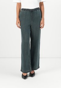 Bukser i denim med vid ben i mørkegrøn, med elastisk talje og subtile syninger ved kanten, parret med sorte spidse sko.