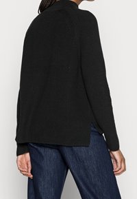 Zwarte geribbelde trui met hoge hals, zijsplitten en lange mouwen. Heeft een gestructureerde stof en een ontspannen pasvorm, gecombineerd met donkere jeans.