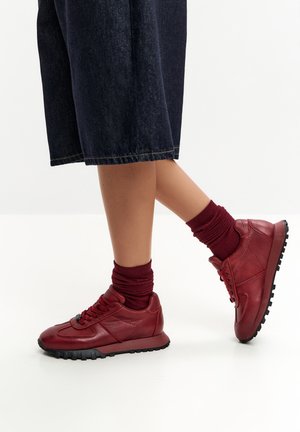 Person iført mørkerøde lædersneakers med matchende sokker og mørke denim vidde bukser på hvid baggrund.