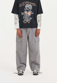 Schwarzes Grafik-T-Shirt mit Totenkopf- und Herzdesign, über einem weißen Langarmshirt getragen. Graue Cargo-Hosen mit Taschen, an den Knöcheln geschnürt.