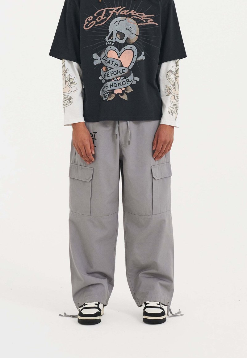 Schwarzes Grafik-T-Shirt mit Totenkopf- und Herzdesign, über einem weißen Langarmshirt getragen. Graue Cargo-Hosen mit Taschen, an den Knöcheln geschnürt.