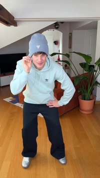 Hele sinine kootud tuukrimüts, heleroheline kapuutsiga dressipluus logoga, mustad laiade säärtega püksid ja hõbedased tennised puidust põrandal, taustal taimed.