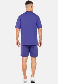 Maglietta viola a maniche corte abbinata a pantaloni corti coordinati, entrambi realizzati in un tessuto leggero e morbido. Include una tasca posteriore sui pantaloni corti.