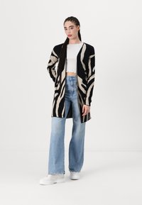 Melnā un bēšā garā jakā ar zebras svītrām, apvienota ar baltu īsā topu un plašām zilām džinsa biksēm, kā arī baltām platformu sporta apaviem.