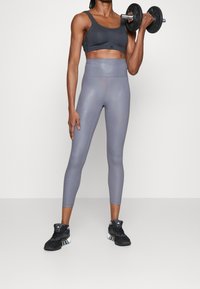 Fit osoba v černém sportovním podprsenku, šedých legínách s vysokým pasem a černých teniskách, drží činku jednou rukou ve výšce ramene.