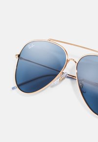 Ochelari de soare aviator cu lentile nuanțate în albastru și un cadru subțire din metal aurit. Prezintă almohade transparente pentru confort și logo-ul mărcii pe lentilă.