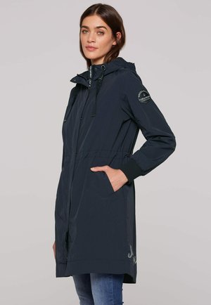 Frau trägt eine marineblaue Kapuzenjacke mit einem Aufnäher am Ärmel und die Hände in den Taschen, steht vor einem einfarbigen hellgrauen Hintergrund.