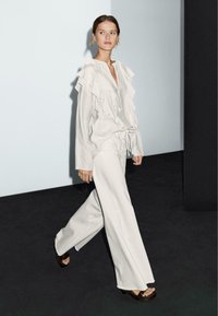 Vrouw wandelt in een losse witte ruffle blouse en wijdvallende witte broek op een donkere vloer met een effen lichte wand als achtergrond.