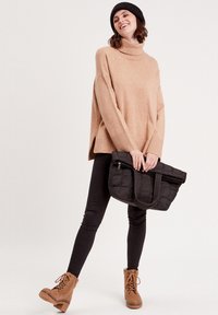 Pull tricot beige oversize à col roulé, associé à un legging noir et des bottes à lacets tan. Tient un sac noir matelassé avec des poignées.