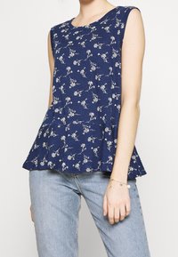 Navy blue sleeveless top with a peplum hem, featuring a floral print in white. Soft material with a smooth texture. Paired with light blue jeans.