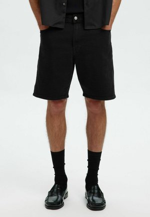Svarta denimshorts, med längd till mitten av låret, med en klassisk femfickdesign, metallknappstängning och fransad fåll. Stylat med svarta strumpor och skor.