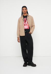 Beige skjorte med knapper over en sort grafik T-shirt med en spiller. Løs sorte bukser og sorte sko fuldender outfittet.