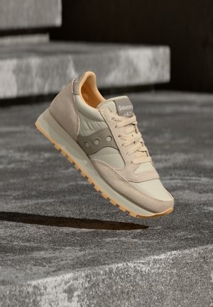Beige and cream Saucony Jazz sneaker with textured sole, floating above gray concrete steps in natural light.