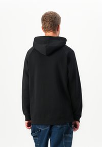 Man wearing a black hooded sweatshirt with long sleeves and blue cargo jeans, shown from the back against a plain white background.