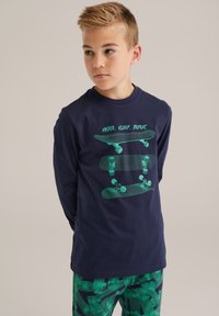 Marinblus med lång �ärm som har en grön skateboardgrafik med texten "SKATE, SLEEP, REPEAT." Matchad med byxor med grönt mönster.