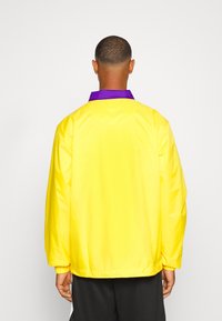 Hombre de espaldas vistiendo una chaqueta amarilla brillante con un collar púrpura y pantalones negros, contra un fondo blanco liso.