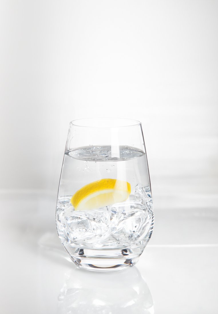 Un vaso de cristal transparente con base redondeada que contiene cubos de hielo y agua, coronado con una rodaja de limón amarillo. El fondo es suave y de colores claros.