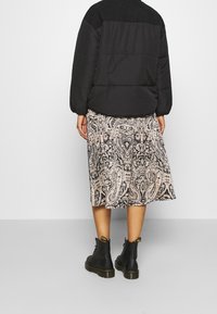 Personne portant une doudoune noire, une jupe midi à motif cachemire, et des bottes noires à lacets, debout devant un fond clair uni.