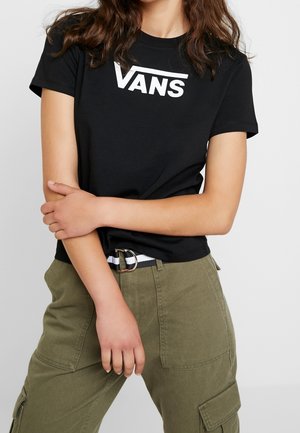 Persona que lleva una camiseta negra de Vans y pantalones cargo verde oliva, con los brazos cruzados sobre la cintura, de pie frente a un fondo liso.