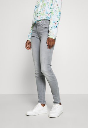 Personne portant un jean skinny gris clair, des baskets blanches et une chemise à manches longues à motif floral, se tenant devant un fond uni.